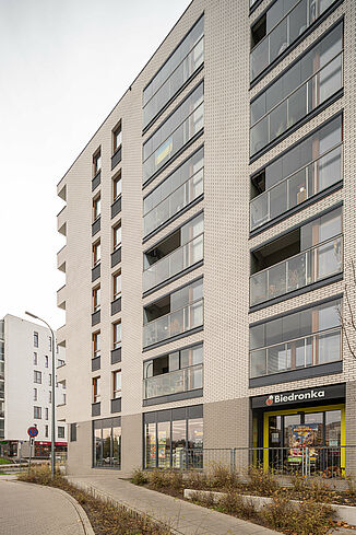 Seitenansicht eines Gebäudes in Warschau mit Supermarkt im Erdgeschoss und darüberliegenden Wohnungen.