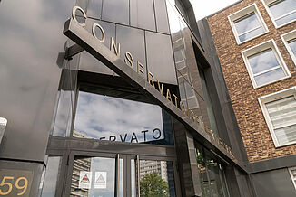 Eingang zu einem modernen Gebäude mit einer schwarzen Fassade, in dem sich die Musikschule Léo Délibes in Paris befindet.