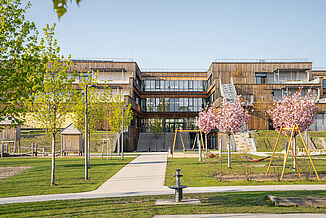 Außenansicht mit Holzfassade, einem Spielplatz, Grünflächen und Treppen die zu den Terrassen des Bildungscampus in Wien führen.